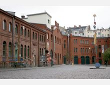 Ziegelrot verkleidete historische Fabrikgebäude mit Rundbogenfenstern, heute als Kultur- und Veranstaltungsräume genutzt, umgeben einen gepflasterten Hof. Im Vordergrund ist eine bunte Skulptur aufgestellt, im Hintergrund ergänzen modernere Wohngebäude die Szene.