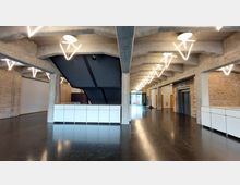 Industrieähnlicher Innenraum mit freiliegenden Ziegelwänden, dunklem Boden und Betonträgern im Deckenbereich. Moderne, geometrische Hängelampen beleuchten die Halle, die einen schwarzen Treppenaufgang, weiße Theken und einen Aufzug umfasst.