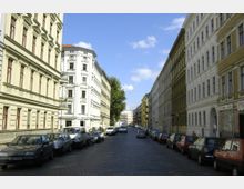 Straßenansicht einer gepflasterten Wohnstraße mit historischen mehrstöckigen Gebäuden in hellen Farben auf beiden Seiten. Entlang der Straße stehen geparkte Autos, und im Hintergrund sind Bäume und weitere Gebäude zu sehen.