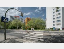 Straßenszene mit Zebrastreifen und einem Verkehrsschild für Fußgängerüberwege im Vordergrund. Im Hintergrund stehen moderne Wohngebäude, darunter ein auffällig orangefarbenes Hochhaus, umgeben von Bäumen und Grünflächen.