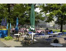 Außenbereich eines Cafés mit mehreren Tischen und Stühlen unter schattenspendenden Bäumen, teilweise mit geschlossenen Sonnenschirmen. Im Hintergrund sind eine Wiese, ein Zaun und moderne Gebäude erkennbar.