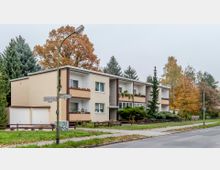 Wohngebäude in einer Wohngegend mit zwei Stockwerken, Balkonen und hellbeiger Fassade, umgeben von Bäumen und Pflanzen im herbstlichen Farbton. Im Vordergrund führt eine Gehwegkreuzung mit Straßenschildern, Straßenlaternen und einem kleinen Grünstreifen vorbei.