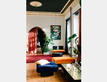 Ein stilvoller Wohnraum mit Holzboden, farbigen Samtmöbeln in Blau und Orange, großen Zimmerpflanzen und modernen Kunstwerken an den Wänden. Eine gewölbte Tür mit roten Vorhängen und ein schwarzes Deckenfries mit Ornamenten betonen den Raum.