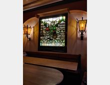 Buntglasfenster mit landschaftlicher Darstellung und einem Mann, der Bier trinkt, eingerahmt von zwei antiken Wandleuchten in einem rustikalen Raum mit Holztischen und Bänken.