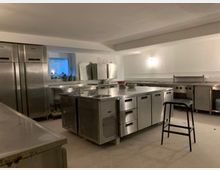 Gewerbliche Küche mit Edelstahlmöbeln, inklusive großer Kühlschränke, Arbeitsflächen und Kochgeräten. Die Beleuchtung ist minimal, und der Raum hat einen gefliesten Boden sowie eine schlichte weiße Wandgestaltung.