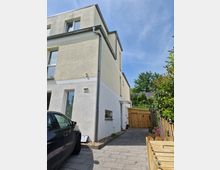 Doppelhaushälfte mit hellgelber und weißer Fassade, großen Fenstern und einem gepflasterten Weg, der zu einem hölzernen Gartentor führt. Im Vordergrund steht ein schwarzes Auto, daneben kleine Beete und Holzzaun mit Pflanzen.