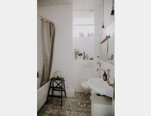 Ein kleines Badezimmer mit weißen Wänden und gemusterten Fliesen auf dem Boden. Es gibt eine Badewanne mit Duschvorhang, ein Waschbecken mit goldener Armatur, eine Wandtoilette und ein kleines Fenster mit einem Rollladen. Ein schwarzer Hocker mit einer Glaskugel steht neben der Badewanne.