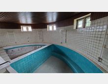 Innenansicht eines leeren, gefliesten Pools mit türkisfarbenen Kacheln und umliegenden weißen Wänden, die mit kleinen Quadratkacheln verkleidet sind. Oben sind kleine Fenster und eine Holzdecke sichtbar.