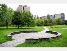 Städtischer Park mit gepflastertem Rundweg und halbrundem Sitzbereich im Vordergrund, umgeben von Rasenflächen, Bäumen und Parkbänken; im Hintergrund Wohngebäude mit mehreren Etagen.