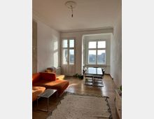 Wohnzimmer mit Holzdielenboden, einem orangefarbenen Sofa, einem kleinen Glastisch und einem hellen Teppich im Vordergrund. Im Hintergrund ein Esstisch mit Stühlen an großen, hohen Fenstern, die eine Aussicht auf Dächer bieten, sowie ein einfacher Wandleuchter an der Decke.