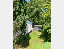 Schmaler, grasbewachsener Gartenweg neben einem weißen Gebäude, links von hohen Nadelgehölzen und rechts von dichtem Laub und Sträuchern gesäumt. Am Ende steht ein kleines graues Gartenhaus vor hohen Bäumen im Hintergrund.