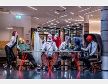 Eine moderne Büroeinrichtung mit einer Gruppe von Personen in auffälligen, bunten Kostümen, die an einem langen Tisch sitzen und stehen. Im Hintergrund sind Säulen, Glaswände und eine helle Deckenbeleuchtung zu sehen.