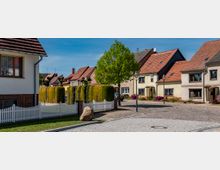 Eine ruhige Wohnstraße mit gepflastertem Gehweg und Reihenhäusern mit rotbraunen Dächern. Im Vordergrund ein weißer Holzzaun, gepflegte Hecken, ein großer Stein und ein einzelner Baum; im Hintergrund klare, blaue Himmel.
