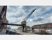 Historischer Hafenbereich mit Backsteingebäuden, einem alten Metallkran und einem Wasserkanal, an dem mehrere Boote festgemacht sind. Im Hintergrund befinden sich weiße Tanks, und der Himmel ist bewölkt.