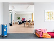 Offener, moderner Wohnraum mit Holzboden, weißen Wänden und einer hohen Decke mit sichtbaren Balken. Im Vordergrund stehen bunte Sitzkissen, im Hintergrund befinden sich blaue Sofas, ein großer Holztisch mit Stühlen und eine Zimmerpflanze; rechteckige, farbige Kunstwerke lehnen an der Wand.