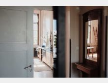 Geöffnete Tür mit Blick in eine helle Küche mit hölzernen Schränken, großen Fenstern und hängenden Lampen; rechts im Bild ein Spiegel, der einen anderen Raum mit einem Tisch und einer Leiter reflektiert.