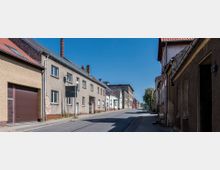 Straße in einer kleinen Stadt mit einfachen, zweigeschossigen Wohnhäusern aus Putz und Ziegeln. Die Straße führt leicht bergauf, ist asphaltiert und von breiten Gehwegen gesäumt, bei klarem Himmel und sommerlichem Wetter.