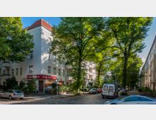 Eine ruhige Straßenkreuzung in einem Wohngebiet mit mehrstöckigen Häusern im klassischen Baustil und roten Dächern, teils von Bäumen beschattet. Im Erdgeschoss eines Gebäudes befindet sich ein Lokal mit dem Schild „Schützen Eck“, umgeben von parkenden Autos und Grünflächen.