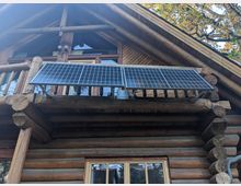 Blockhütte aus Holzstämmen mit einem Balkon, auf dem zwei Solarpaneele montiert sind; darüber ein Giebeldach mit großen Fenstern. Im Hintergrund Äste eines Baumes sichtbar.