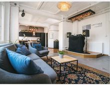Wohnraum in modernem Loft-Stil mit Sitzbereich bestehend aus grauen Sofas, blauen Kissen und einem Holztisch auf gemustertem Teppich. Im Hintergrund befinden sich eine offene Küche mit schwarzen Schränken und ein erhöhtes Podest mit DJ-Pult vor einer weißen Wand mit dem Schriftzug "Loftyard Studios".
