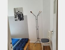 Eckiger Raum mit Holzboden, einem Metall-Kleiderständer, einem weißen Stuhl und einem Holzbett mit blauer Bettwäsche. An der Wand hängt ein Schwarz-Weiß-Poster mit der Aufschrift "PARIS".