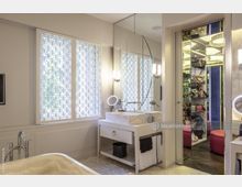 Modernes Badezimmer mit hellem Waschtisch aus Stein, ovalem Wandspiegel und Leuchten an beiden Seiten. Große Fenster mit geometrischen Mustermotiven, ein freistehender Badewannan-Rand im Vordergrund und ein Spiegel-Schrank mit Taschen auf der rechten Seite.