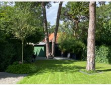 Garten mit gepflegtem Rasen, hohen Kiefern und dichtem Laubwerk. Im Hintergrund sind eine grüne Holzwand und ein Dach mit roten Ziegeln zu sehen, umgeben von Hecken und weiteren Bäumen.