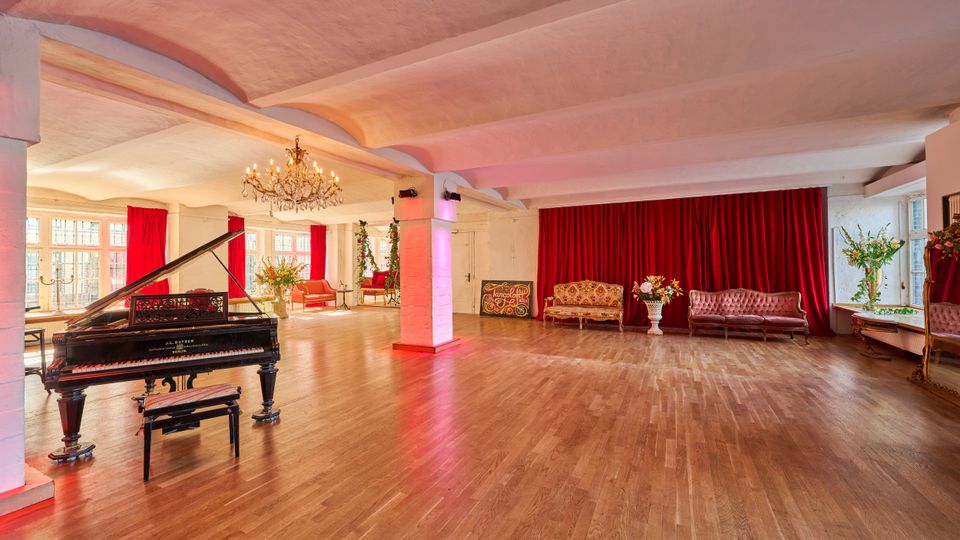 Großer Veranstaltungsraum mit Holzparkett, weißer Decke mit sichtbaren Trägern und Kronleuchtern sowie hohen Fenstern mit roten Vorhängen. Links steht ein Flügel, rechts bilden eine rote Bühnenvorhangwand, antike Sofas und Blumenarrangements eine Lounge- und Bühnenzone.
