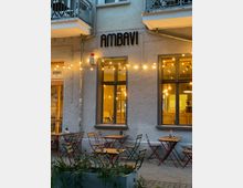 Außenbereich eines Cafés mit dem Namen "AMBAVI" auf einer grauen Gebäudefassade. Vor dem Eingang stehen rote und grüne Metalltische mit Holzstühlen, umgeben von Lichterketten und einem kleinen Blumenbeet.