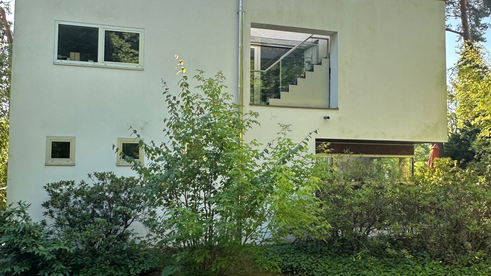 Weißes, kubisches Wohnhaus mit Flachdach und rechteckigen Fenstern, teilweise auf Stützen erhöht, mit einem großen quadratischen Ausschnitt, der eine innenliegende Treppe zeigt. Davor liegt ein Garten mit Rasen, Sträuchern und einem kleinen Baum, umgeben von hohen Kiefern.
