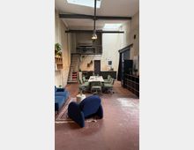 Loftartiger Raum mit hoher Decke, minimalistischer Einrichtung und Industriestil-Elementen. Zentral ein großer Besprechungstisch mit grünen Bürostühlen, eine Metalltreppe führt zu einer offenen Zwischengalerie, und im Vordergrund stehen moderne blaue Sessel auf einem Teppich.