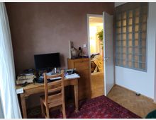 Ein Raum mit einem Schreibtisch aus Holz, darauf ein Monitor, Bücher und Kabel. Rechts ist eine geöffnete Tür, die in einen anderen, hell beleuchteten Raum führt. Daneben befindet sich eine Wand aus Glasbausteinen. Der Boden besteht aus Holzdielen und ein roter Teppich liegt teilweise sichtbar am unteren Bildrand.