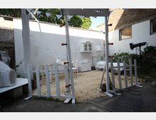 Ein kleiner Innenhof mit weißen, rustikalen Möbeln, darunter ein Bett, ein Tisch und ein Schrank, eingerichtet in einer sandbedeckten Fläche. Die Szenerie ist von weißen Wänden umgeben, dekoriert mit Girlanden, einem kleinen Holzzaun und umgeben von Pflanzen und Backsteinmauern.