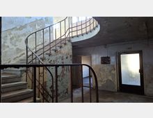 Innenraum eines heruntergekommenen Treppenhauses mit geschwungener Steintreppe und rostigem Metallgeländer, die zu einer oberen Galerie führt. Abgeplatzter Putz an den Wänden und eine Tür mit Milchglasfenster prägen die karge, ältere Gebäudesituation.