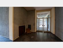 Ein leerer Innenraum mit grauen Wänden und schwarzem Marmorboden. Vorne rechts führt eine doppelflügelige Glastür zu einem hellen, gewölbten Flur mit Fensterelementen; links steht ein gepolsterter brauner Stuhl vor einem Heizkörper.