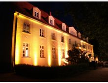 Beleuchtete Fassade eines großen, mehrstöckigen Gebäudes mit gelber Außenbeleuchtung in der Nacht, einem roten Schrägdach und mehreren Dachgauben. Im Vordergrund sind Bäume und Büsche zu sehen.