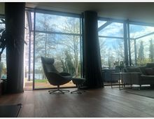 Wohnzimmer mit großen bodentiefen Fenstern, die den Blick auf einen Garten mit Bäumen und einem See im Hintergrund freigeben. Im Raum befinden sich ein moderner Sessel mit Hocker, ein Sofa, ein kleiner Beistelltisch mit einer Vase sowie ein Holzboden und dunkle Vorhänge.