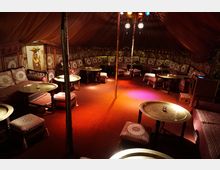 Innenraum eines orientalischen Zeltes mit niedrigen Tischen aus goldenem Metall, Sitzpolstern mit gemusterten Bezügen und warmem, gedämpftem Licht. Die Wände und Decke sind mit Stoff bespannt, und an einer Wand hängt ein dekoratives Gemälde.