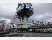 Futuristisches Gebäude in Form eines Baums mit bunten, würfelförmigen Elementen, das auf einem schmalen Stützpfeiler ruht. Im Vordergrund eine belebte Straßenkreuzung mit Autos, Radfahrern und Fußgängern sowie einer Brücke mit der Aufschrift "HERTHA BSC".