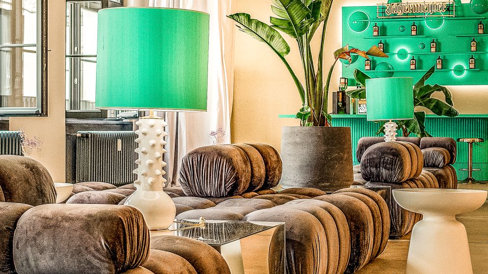 Modern gestaltete Lounge mit modularen, braunen Samtsofas, niedrigen Beistelltischen und großen grünen Tischlampen. Im Hintergrund stehen hohe Topfpflanzen vor einer grünen Barwand, dazu hohe Decken mit Sternleuchten und hohe Fenster mit Vorhängen.