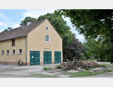 Gelbes landwirtschaftliches Gebäude mit grün gestrichenen Toren und einem Ziegelsockel, umgeben von Bäumen. Im Vordergrund befinden sich ein Schubkarren und ein Haufen Erde neben einem unbefestigten Boden.