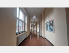 Langer, heller Flur in einem Gebäude mit beigen Wänden, großen Fenstern auf der linken Seite, einer Pinnwand auf der rechten Wand und einer braunen glänzenden Bodenfläche. Hängende Deckenlampen und Heizkörper unter den Fenstern ergänzen die Einrichtung.