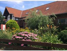 Fachwerkhaus mit rotem Ziegeldach, umgeben von einem bunten Garten mit Blumen und Sträuchern. Im Vordergrund steht ein Holzzaun, unter einem blauen Himmel.
