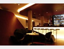 Elegante Bar mit einer beleuchteten Theke, hohen Hockern und einer Rückwand voller Flaschen. Im Vordergrund befindet sich eine Sitzecke mit dunklen Sesseln, einem kleinen runden Tisch und dezenten Dekorationen.