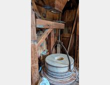 Innenraum einer historischen Mühle mit hölzerner Balkenkonstruktion und grob vertäfelten Wänden. Im Vordergrund steht ein großer runder Mühlstein mit zentraler Öffnung und Metallbügel, daneben sind alte Zahnräder und mechanische Bauteile sichtbar.