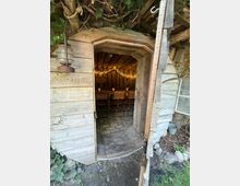 Blick durch eine offene Holztür in eine kleine, rustikale Holzhütte mit Tonnenform, Holzbalkendecke und Steinboden. Im Inneren stehen Holzklappstühle, dekorative Laternen und eine Lichterkette; außen sind Holzverschalung, Efeu und ein altes Lampengehäuse zu sehen.