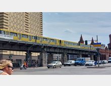 Gelbe U-Bahn der Berliner Verkehrsbetriebe (BVG) auf einer erhöhten Stahlbrücke vor einer Backsteinfassade mit Rautenmustern; darunter Autos, Radfahrer und Fußgänger an einer belebten Straßenkreuzung, im Hintergrund die markanten Türme der Oberbaumbrücke.