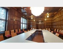 Holzvertäfelter Konferenzraum mit rechteckig angeordneten weißen Tischen und umlaufenden Stuhlreihen. Große Buntglasfenster und eine zentrale moderne Hängelampe prägen den Raum, der durch die dunklen Holzdetails eine klassische Atmosphäre erhält.