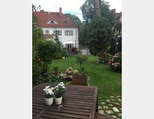 Ein gepflegter Garten mit Rasenfläche, Blumenbeeten und einem Holztisch mit Blumentöpfen im Vordergrund. Im Hintergrund steht ein zweigeschossiges Haus mit roten Dachziegeln, weißen Fensterläden und einer Terrasse.