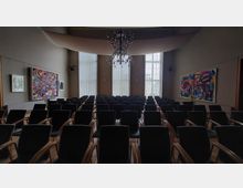 Ein moderner Konferenzraum mit schwarzen Stühlen in mehreren Reihen, hohen Fenstern mit vertikalen Jalousien und bunten abstrakten Gemälden an den Wänden. Eine große Deckenleuchte hängt zentral im Raum.